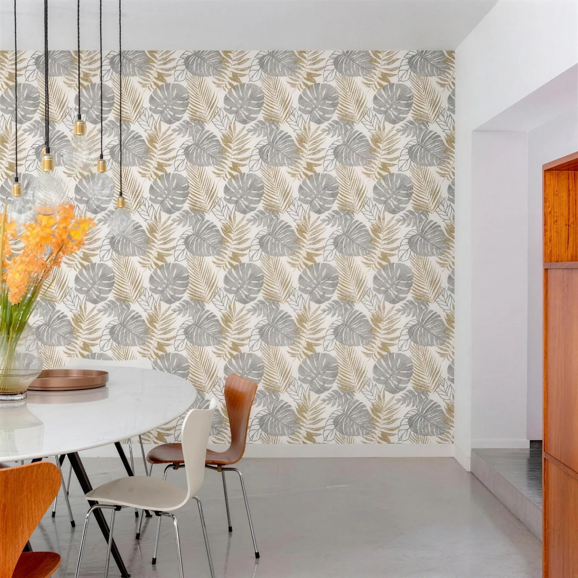 Sala de jantar clara com papel de parede de folhagens em cinza e dourado, mesa redonda branca e cadeiras de madeira.