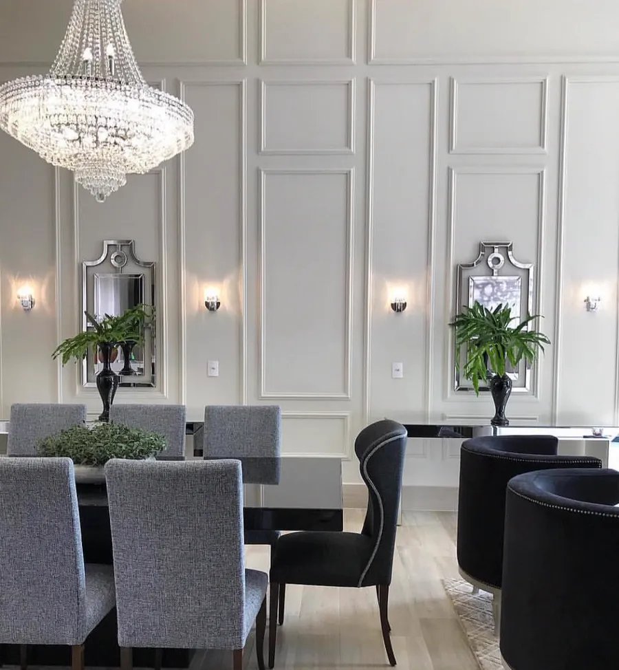 Sala de jantar com parede inteira em boiserie branca, lustre de cristal e cadeiras estofadas cinza.