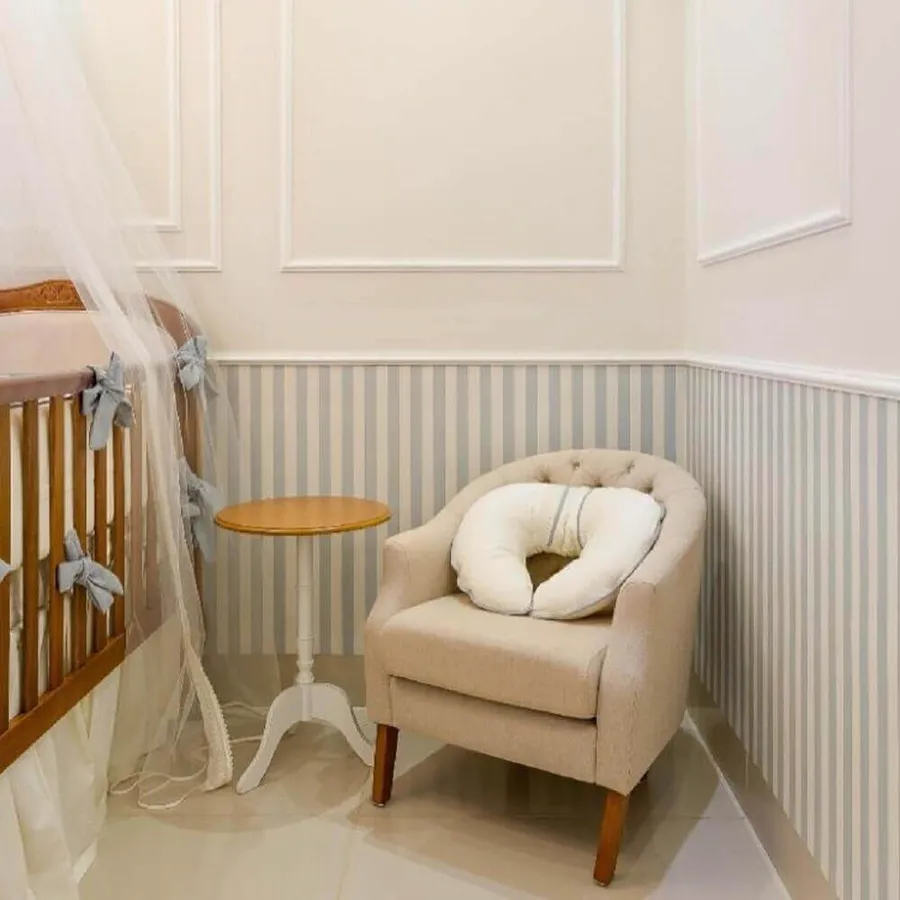Quarto de bebê com boiserie branca acima de listras azul-claras, berço e poltrona bege.