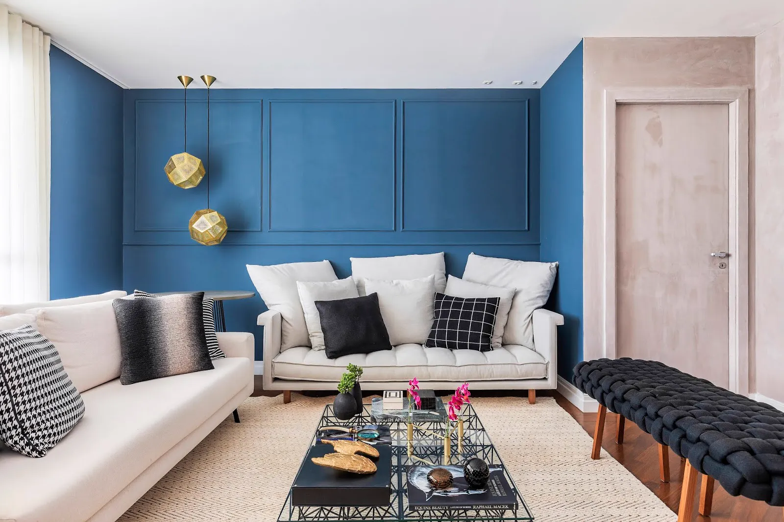 Sala com parede azul em boiserie, pendentes dourados e sofás claros com almofadas pretas e brancas.