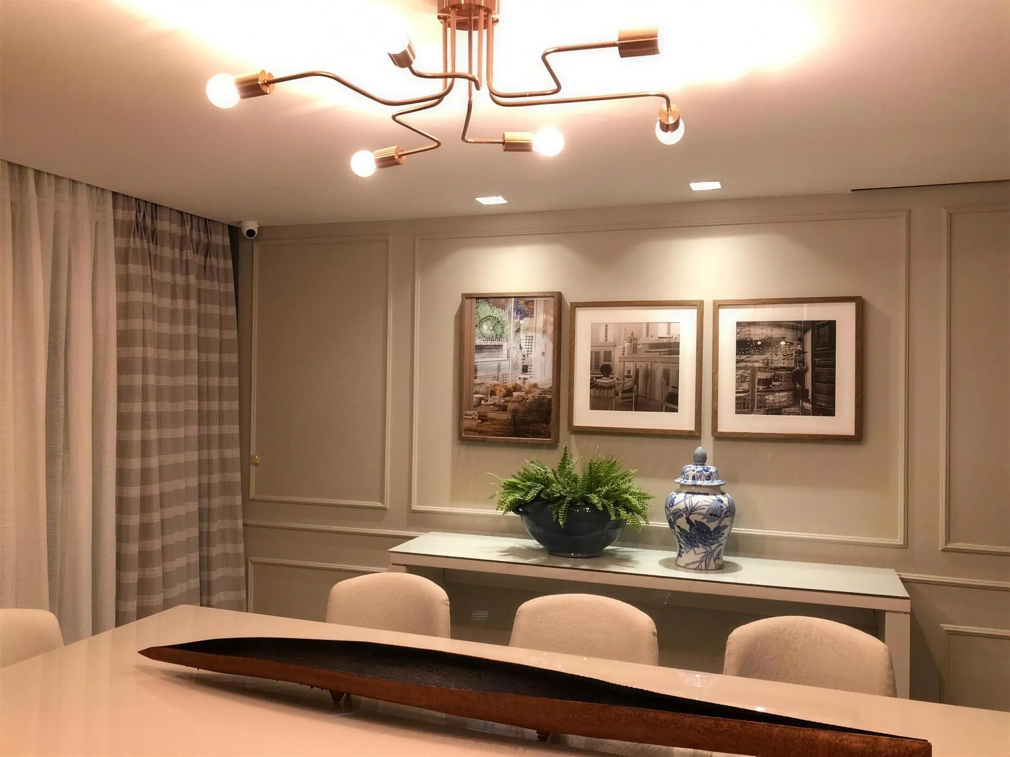 Sala de jantar com molduras de boiserie na parede bege, três quadros e aparador com samambaia.