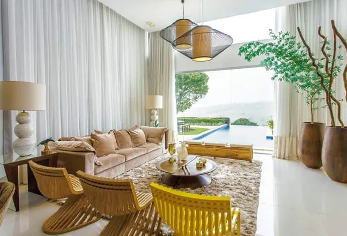 Sala de pé-direito alto com cortinas brancas translúcidas, sofá bege e vista para a piscina.