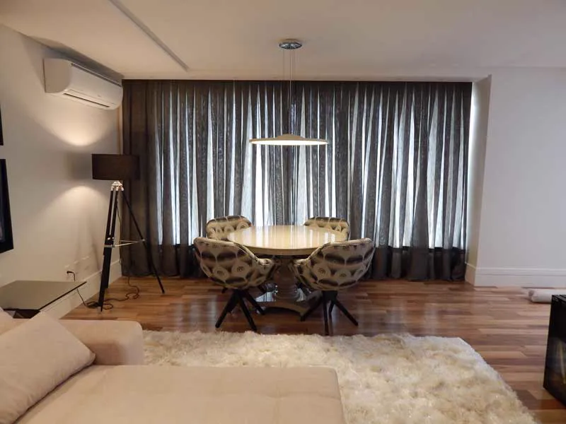 Sala de jantar com cortina cinza pregueada cobrindo toda a parede de janelas e tapete branco felpudo.
