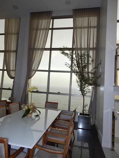 Sala de jantar com cortinas translúcidas drapeadas em janelas altas voltadas para o mar.