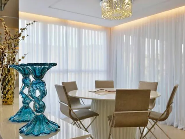 Sala de jantar com cortinas brancas translúcidas cobrindo as janelas e mesa redonda clara.
