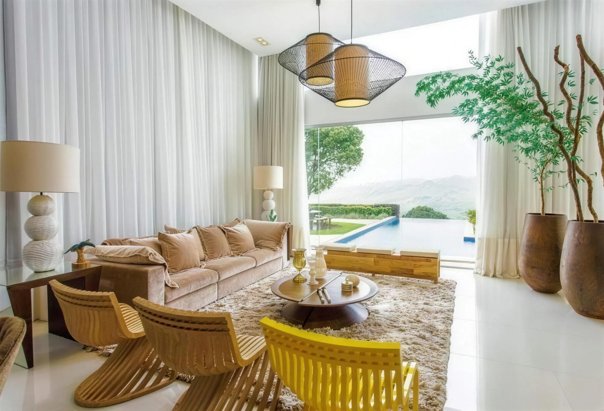 Sala de pé-direito alto com cortinas brancas translúcidas, sofá bege e vista para a piscina.