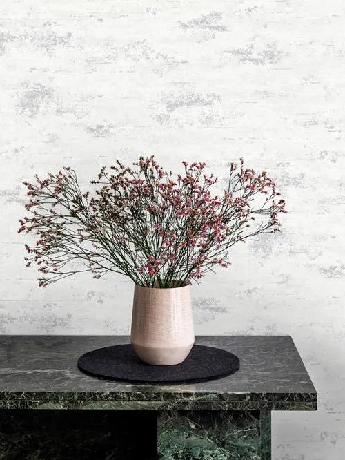 Papel de parede claro com manchas cinza atrás de um aparador de mármore verde com vaso de flores.