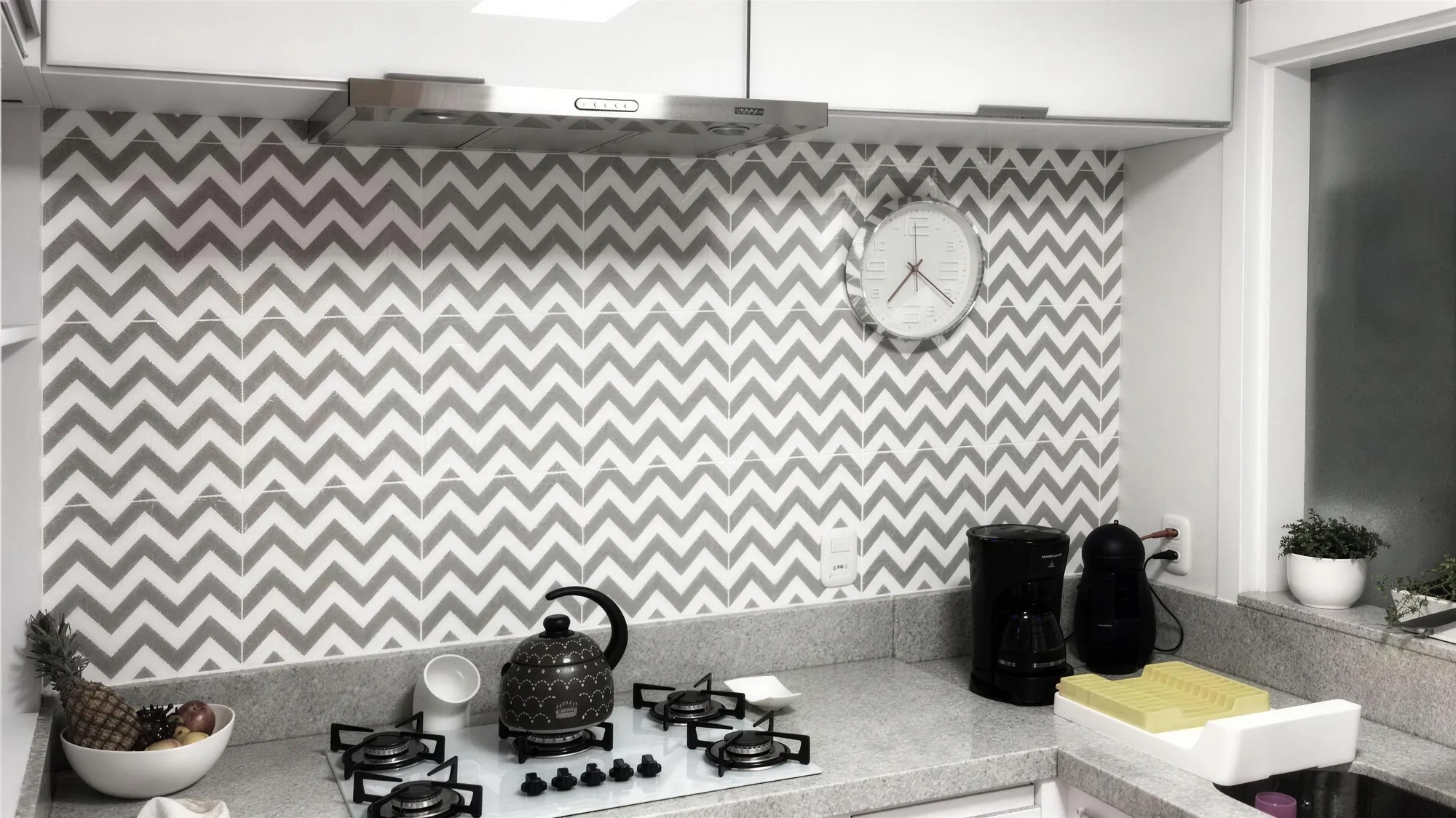 Parede de cozinha com pastilhas em zigue-zague cinza e branco, relógio e cooktop.