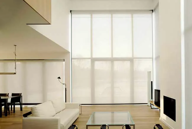 Sala de pé-direito duplo com persianas rolo translúcidas do teto ao chão e sofá branco.
