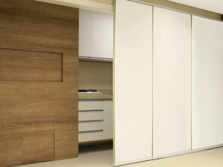 Cortina painel com três folhas brancas deslizantes fechando a passagem para a cozinha, ao lado de um painel de madeira.
