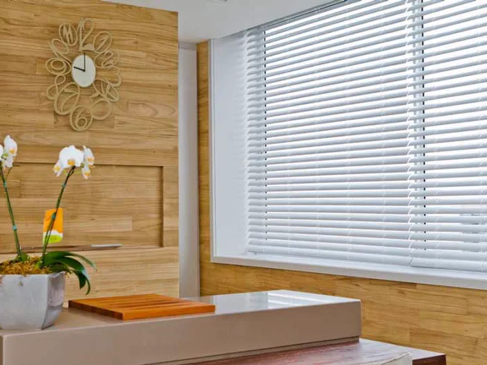 Persiana horizontal de PVC branca numa janela ampla, ao lado de parede de madeira clara e uma orquídea.