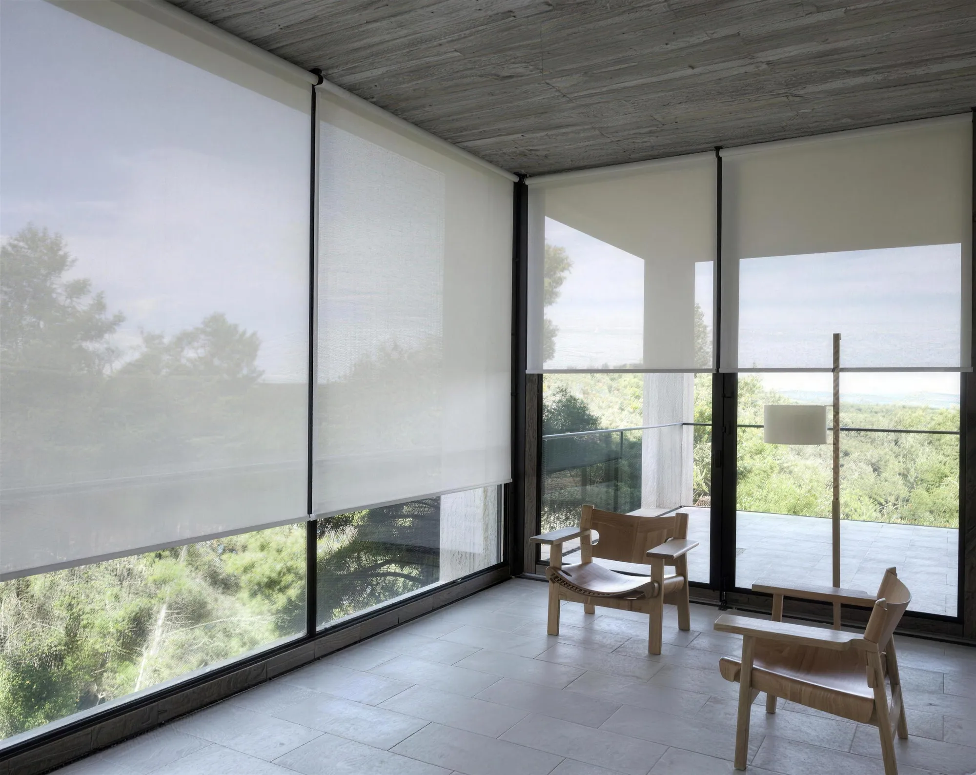 Varanda envidraçada com persianas rolo em tela solar, poltronas de madeira e vista para as árvores.