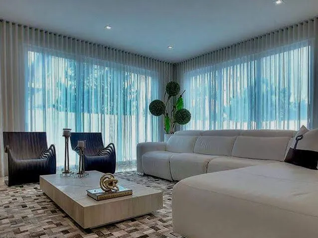 Sala ampla com cortinas translúcidas, sofá claro em L e tapete de couro em mosaico de tons marrons e cinza.