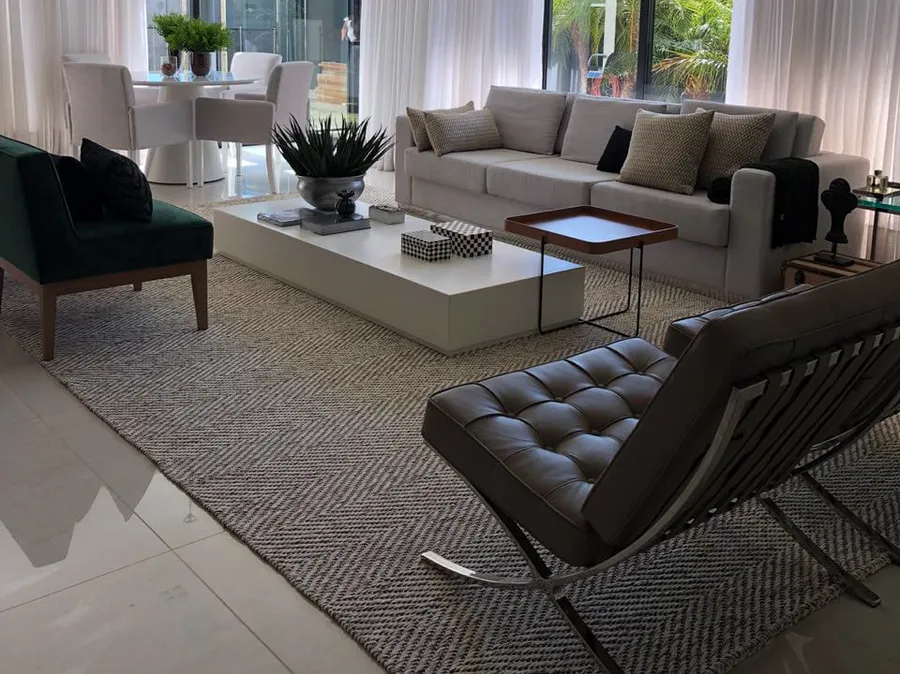 Living claro com cortinas brancas, poltrona de couro e tapete trançado em espinha de peixe cinza e cru.